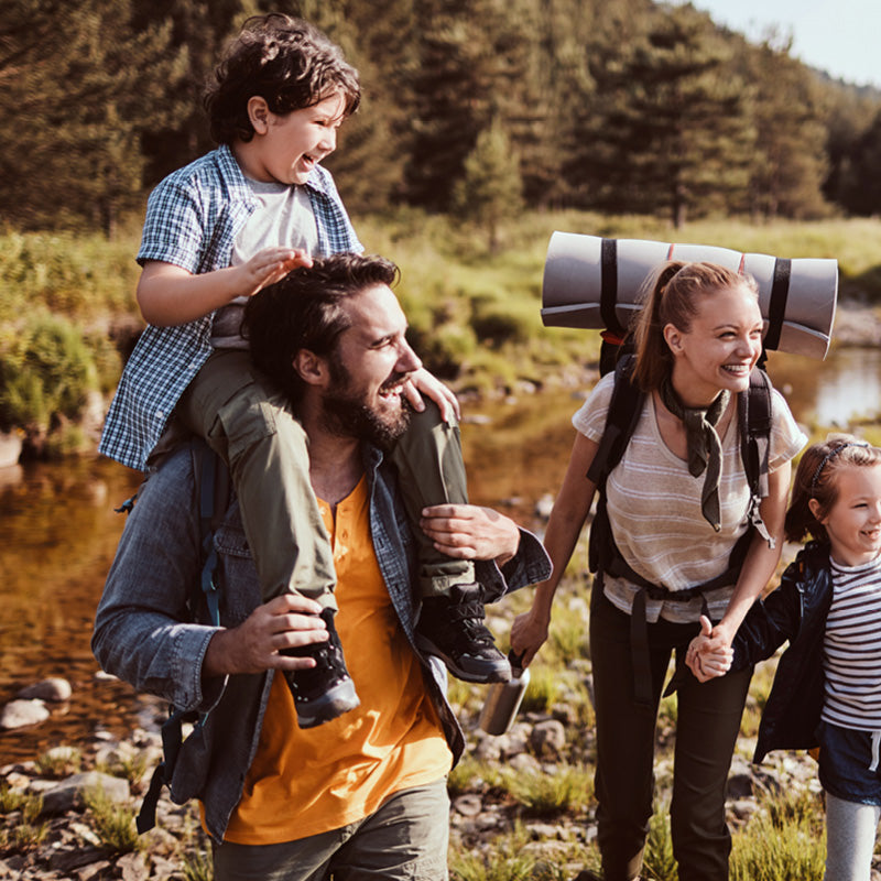 A family of four hiking outdoors near a stream, with a man carrying a child on his shoulders, a woman carrying a sleeping bag on her back, and another child walking beside them. They are dressed in casual outdoor clothing and wearing backpacks.