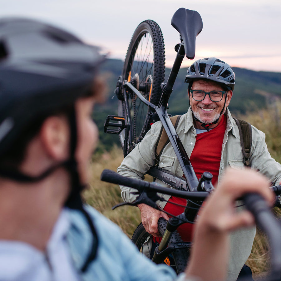 Two mountain bikers outdoors on a grassy hill at dusk, with one person in the foreground in profile and another smiling man holding a bike rack with a mountain bike. Both wear helmets.